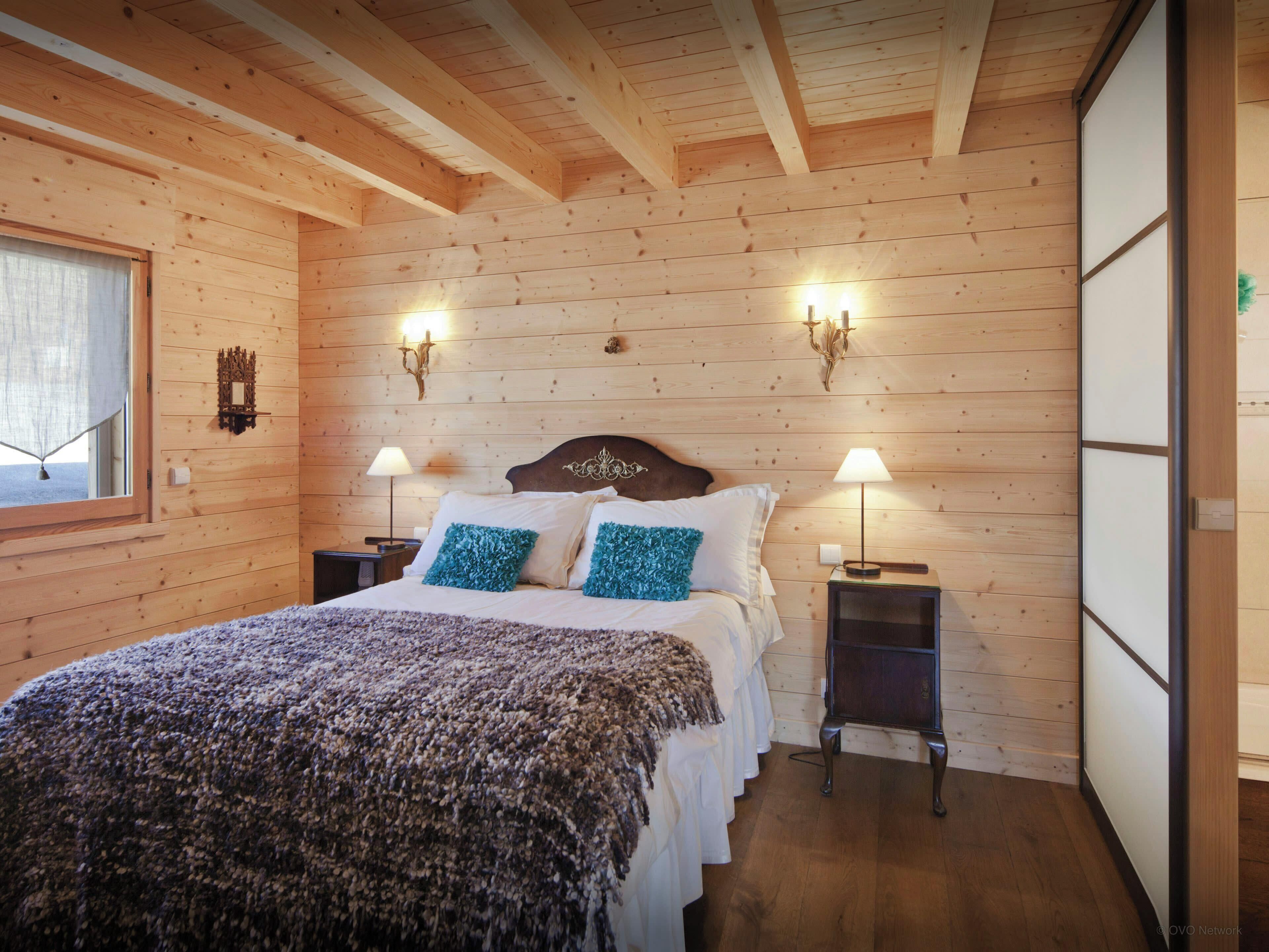 A wooden-paneled bedroom with a bed, two bedside lamps, and a window with curtains.