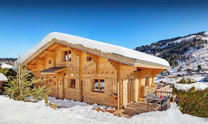 Una cabaña de madera con techo nevado, una terraza de madera y montañas al fondo.