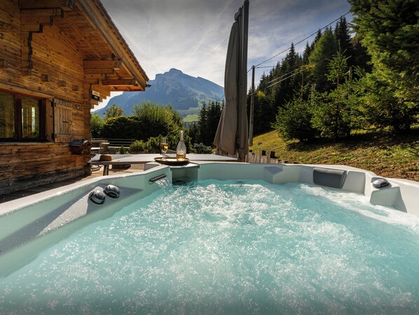 A wooden cabin with a hot tub and outdoor seating, surrounded by a forest and mountains.