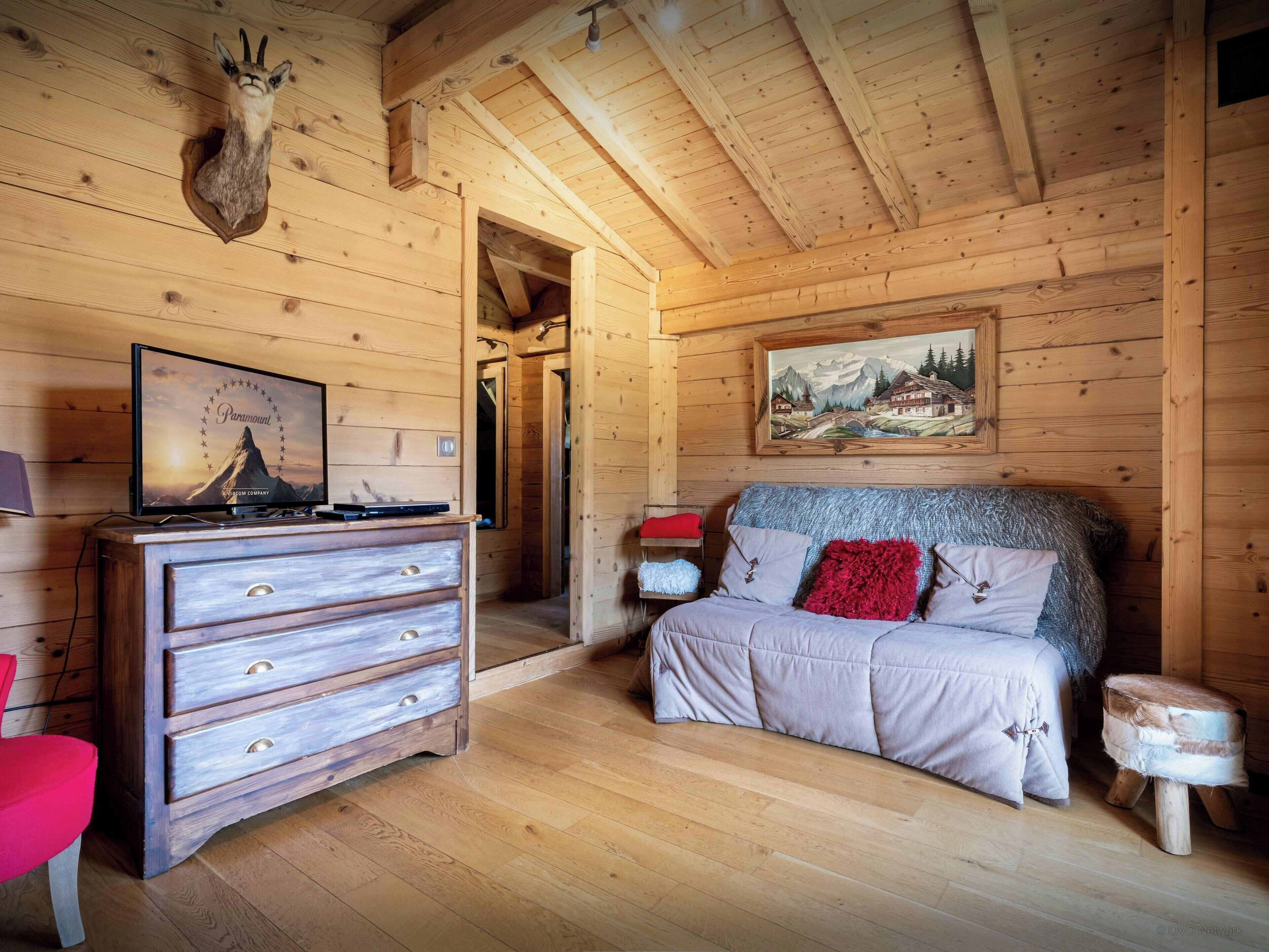 Ein Zimmer in einer Holzhütte mit einem Bett, einem Schminktisch, einem aufgehängten Tierkopf, einem Gemälde und einem Fernseher, der eine Bergbahn zeigt.