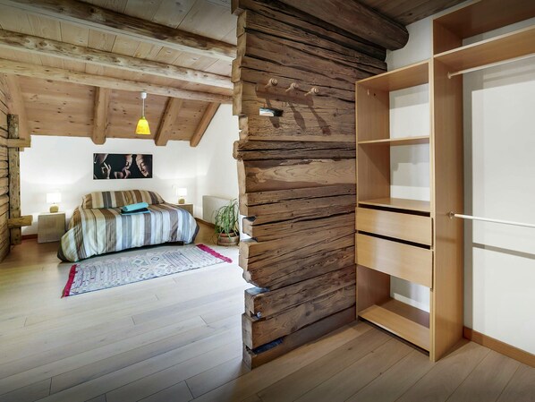 A bedroom with a wooden ceiling, a bed with striped bedding, a bedside lamp, a potted plant, and a wooden wardrobe.