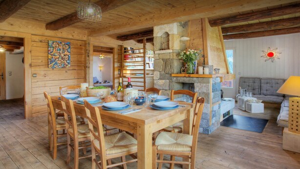 Una mesa de comedor de madera dispuesta para seis personas, con sillas, una chimenea de piedra, una estantería y un rincón de estar acogedor con un sofá y una lámpara.