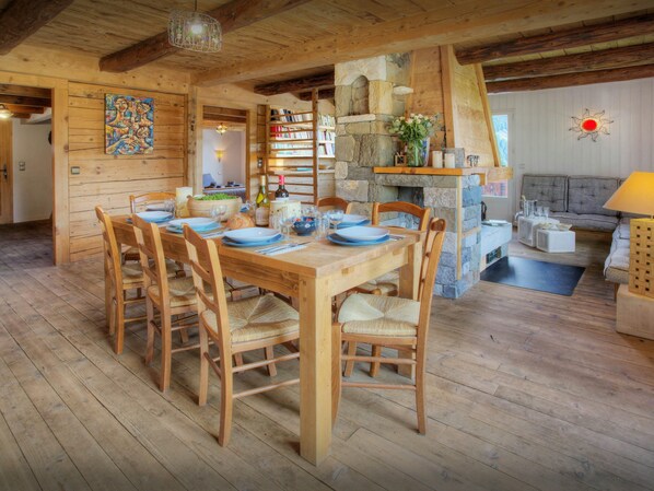 A wooden dining table set for six with chairs, a stone fireplace, a bookshelf, and a cozy seating area with a sofa and a lamp.