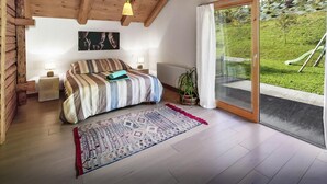 Un dormitorio con cama, mesitas de noche, lámpara colgante, ventana con vistas a la vegetación y puerta que da al exterior.