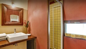 Baño con tocador de madera, lavamanos blanco y espejo encima. Se ve una ducha con azulejos amarillos a través de la puerta.