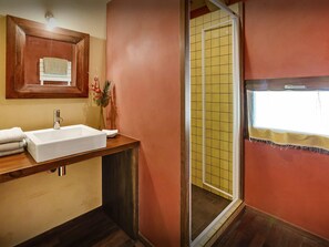 A bathroom with a wooden vanity, a white sink, and a mirror above it. There is a shower with yellow tiles visible through the door.