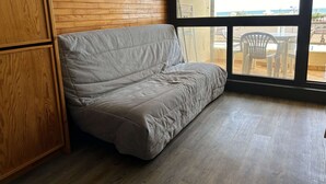 Ein Zimmer mit einem hölzernen Kopfende, einem grauen Sofa und einem großen Fenster mit Blick ins Freie.