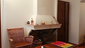 Un coin douillet avec un fauteuil en osier, une petite table sur laquelle est posé un plateau coloré, et un poêle à bois agrémenté d’objets décoratifs.