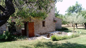 Un bâtiment rustique en pierre, doté d’une porte en bois, d’une petite fenêtre et d’un banc en pierre, situé dans un jardin avec de l’herbe et des arbres.