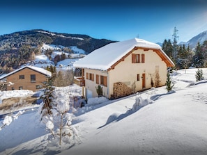 Une maison de montagne recouverte de neige, dotée d’un balcon en bois, d’un mur en pierre et offrant une vue dégagée sur le paysage enneigé environnant.