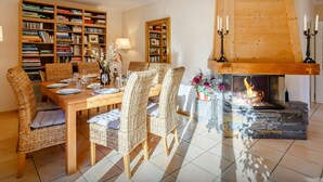 A dining area with a wooden table and wicker chairs, surrounded by bookshelves, a fireplace, and a lamp. - Les Roses des Alpes - luxury mountain break, views & spa - OVO Network (Thônes)