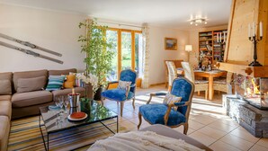 A living room with a sofa, chairs, a coffee table, and a fireplace. There is a bookshelf and a dining area in the background. - Les Roses des Alpes - luxury mountain break, views & spa - OVO Network (Thônes)