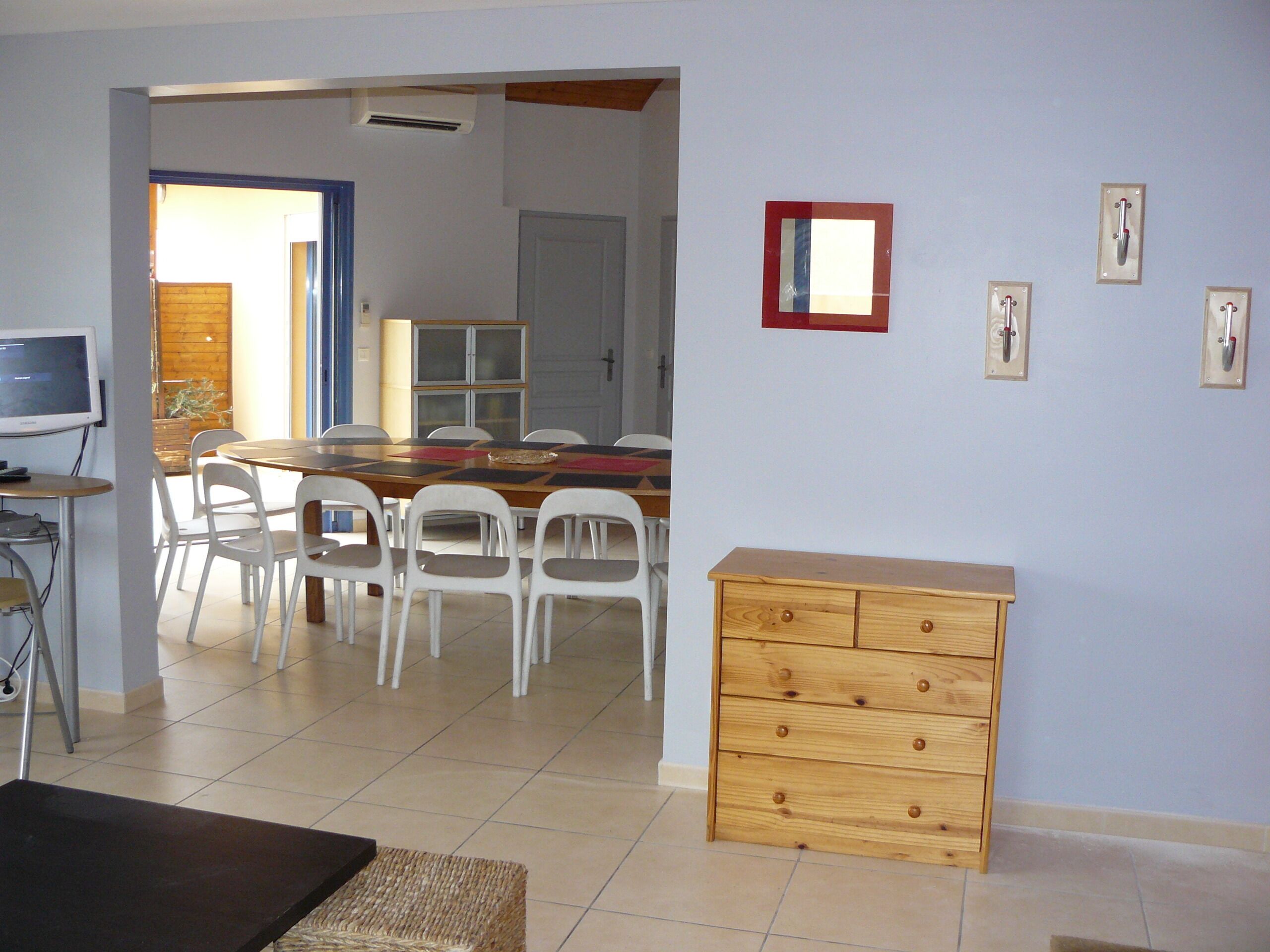 Un comedor con una mesa de madera y sillas blancas, un mueble de madera pequeño y un televisor sobre un soporte.