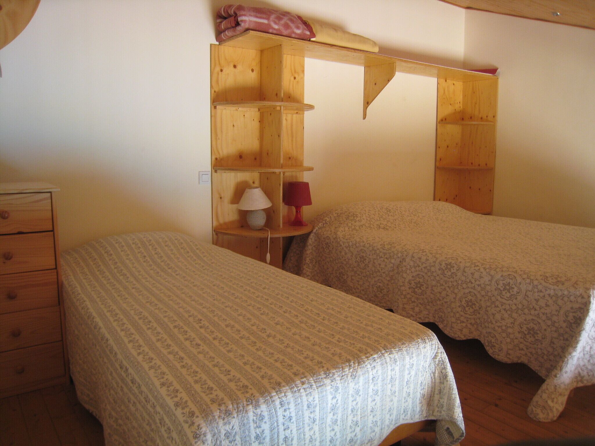 A small, cozy room with two single beds, a wooden dresser, and a shelf unit with a lamp and a red object.