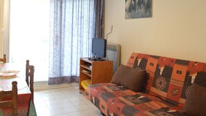 Una sala de estar con sofá, televisor sobre un mueble, mesa de comedor con sillas y un amplio ventanal con cortinas.