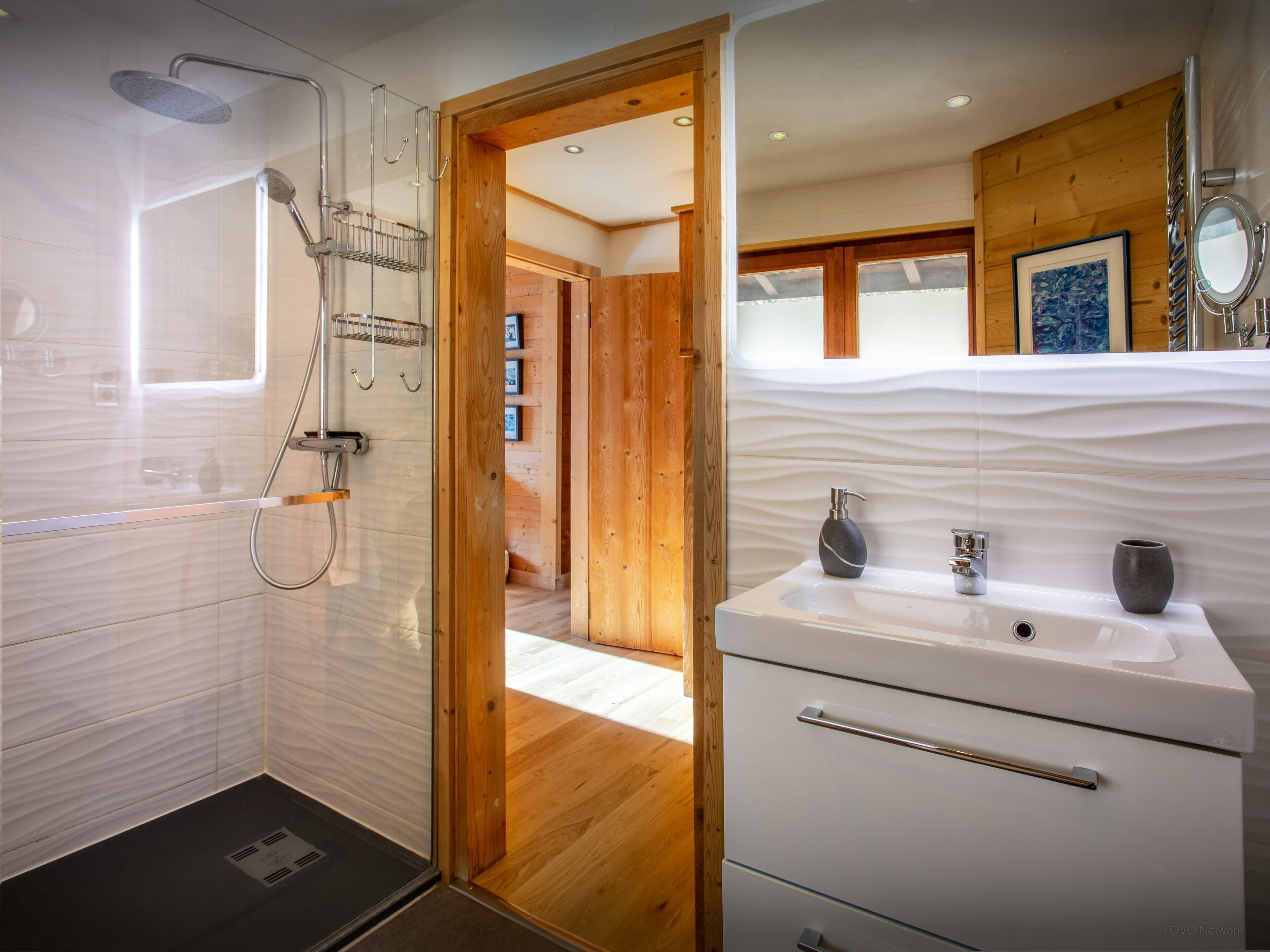 Un baño moderno con ducha de vidrio, lavamanos blanco con lavabo de mármol y detalles de madera.