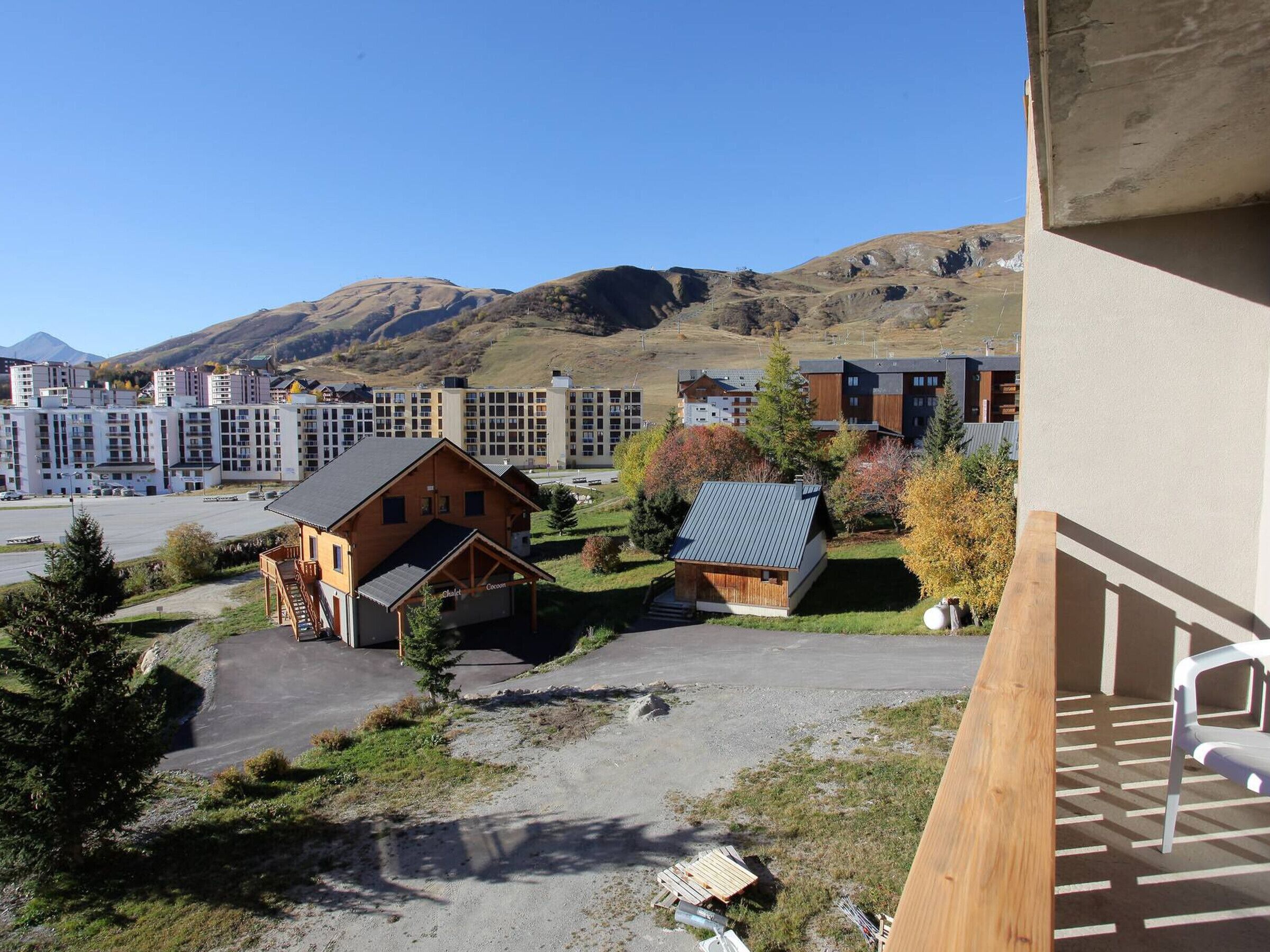 Vista de un hotel con edificio de estilo chalet, un área de estacionamiento y un telón de fondo de edificios modernos y montañas.