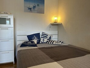 A bedroom with a bed, a dresser, a wall-mounted lamp, and a painting of boats.