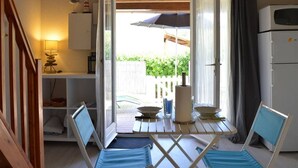 A small, open-plan living space with a dining area, a kitchenette, and a view of a wooden deck and greenery. - Beachside Studio with Mezzanine, Pool, Tennis, 800m to Beach - 4 Guests, Pet-Friendly (Fleury-Saint-Pierre-la-Mer)