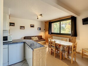 Eine kompakte Küche mit Mikrowelle, kleinem Kühlschrank, einem Holzespeisetisch mit Stühlen und einer Couch. An der Wand befinden sich gerahmte Bilder, ein Deckenventilator und ein Fenster mit Vorhängen.