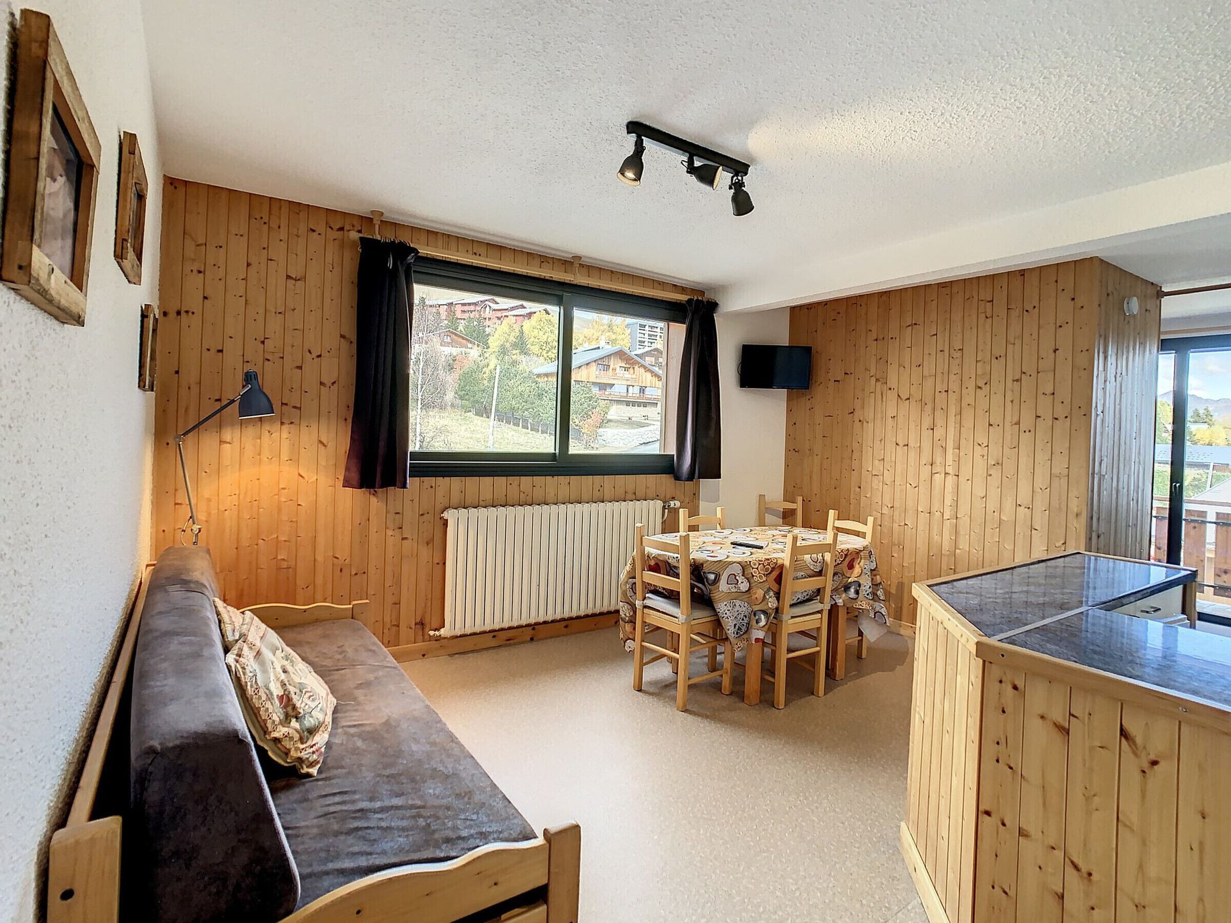 A cozy room with a wooden floor, a sofa, a dining table with chairs, a radiator, a window with curtains, and a television on the wall.