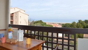 A balcony with a wooden table, a chair, a bottle, a mug, and a coffee maker. The view includes buildings and trees. - Studio with mezzanine for 5, near the beach, pets allowed - Saint Pierre la Mer (Fleury-Saint-Pierre-la-Mer)