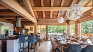 A spacious kitchen with wooden beams, a dining area with a long table and chairs, and large windows offering a view of the outdoors.