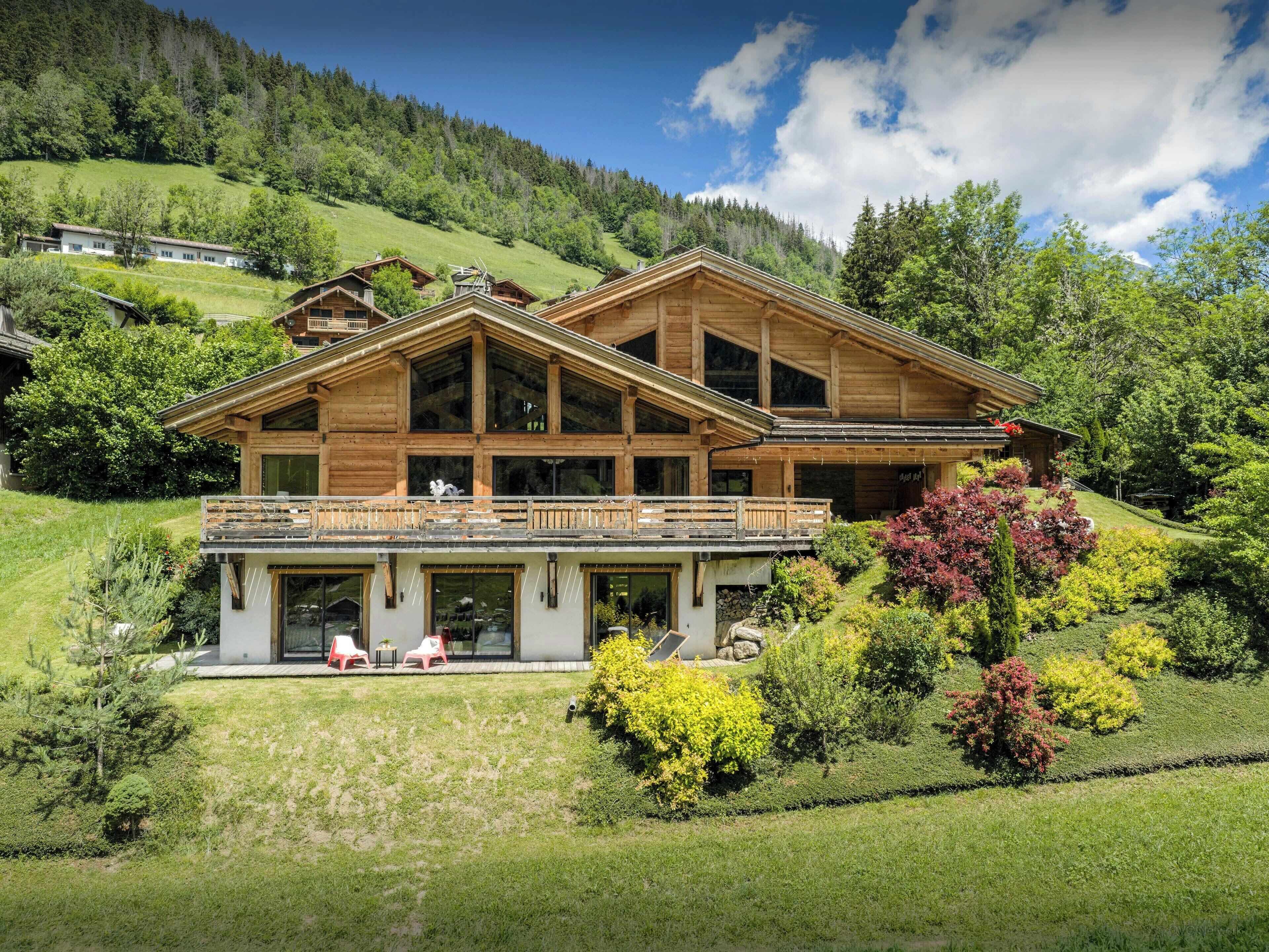 Een ruime houten chalet met balkon, omgeven door weelderige begroeiing en een bergachtig landschap.