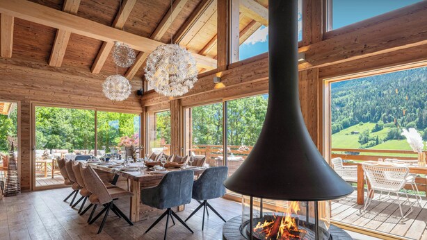 A wooden interior with a fireplace, dining table, chairs, and large windows.