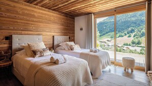 A room with two beds, wooden ceiling, large windows, and a view of a valley.