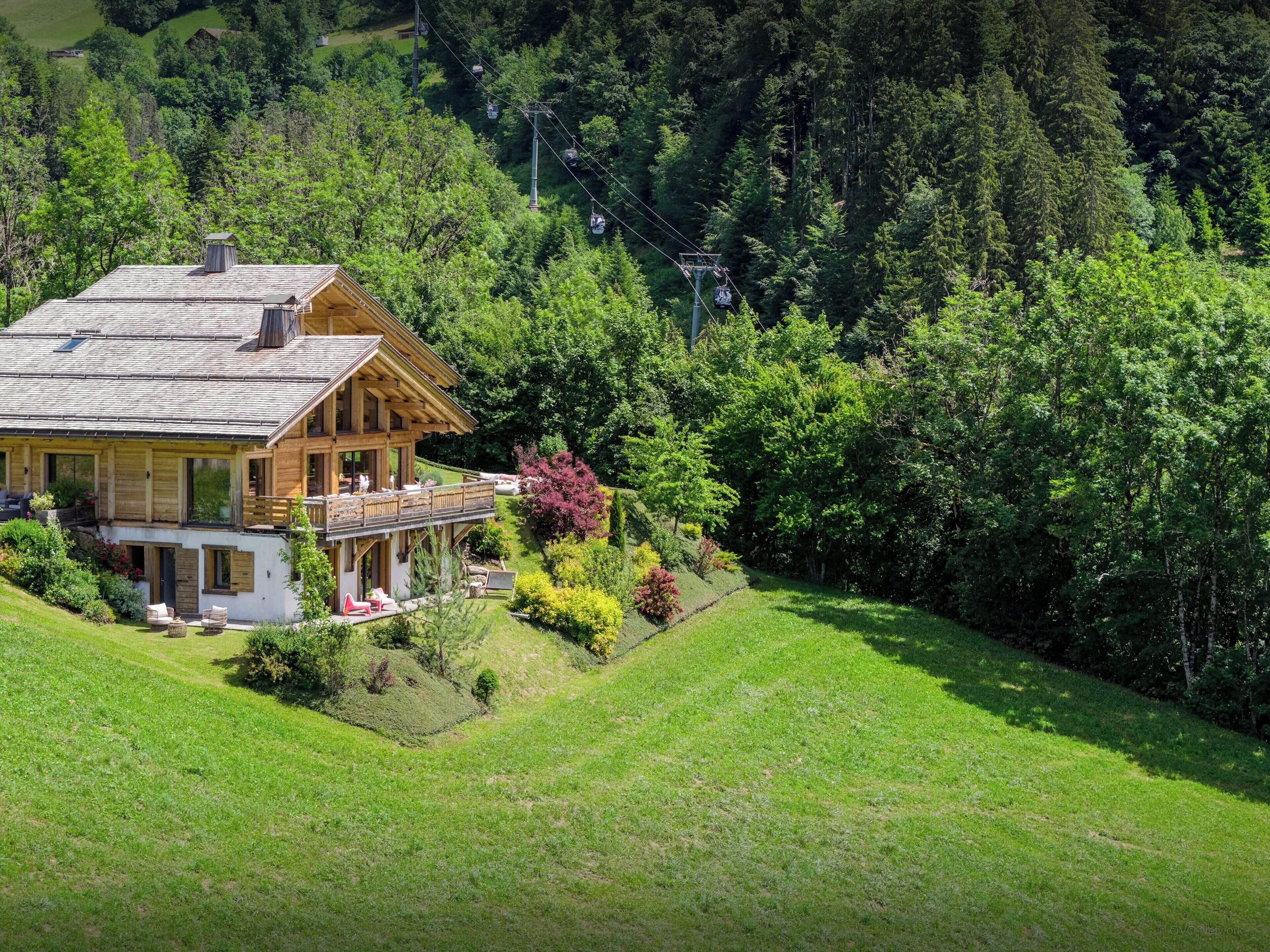 A wooden house with a balcony, surrounded by lush greenery and a ski lift in the background.