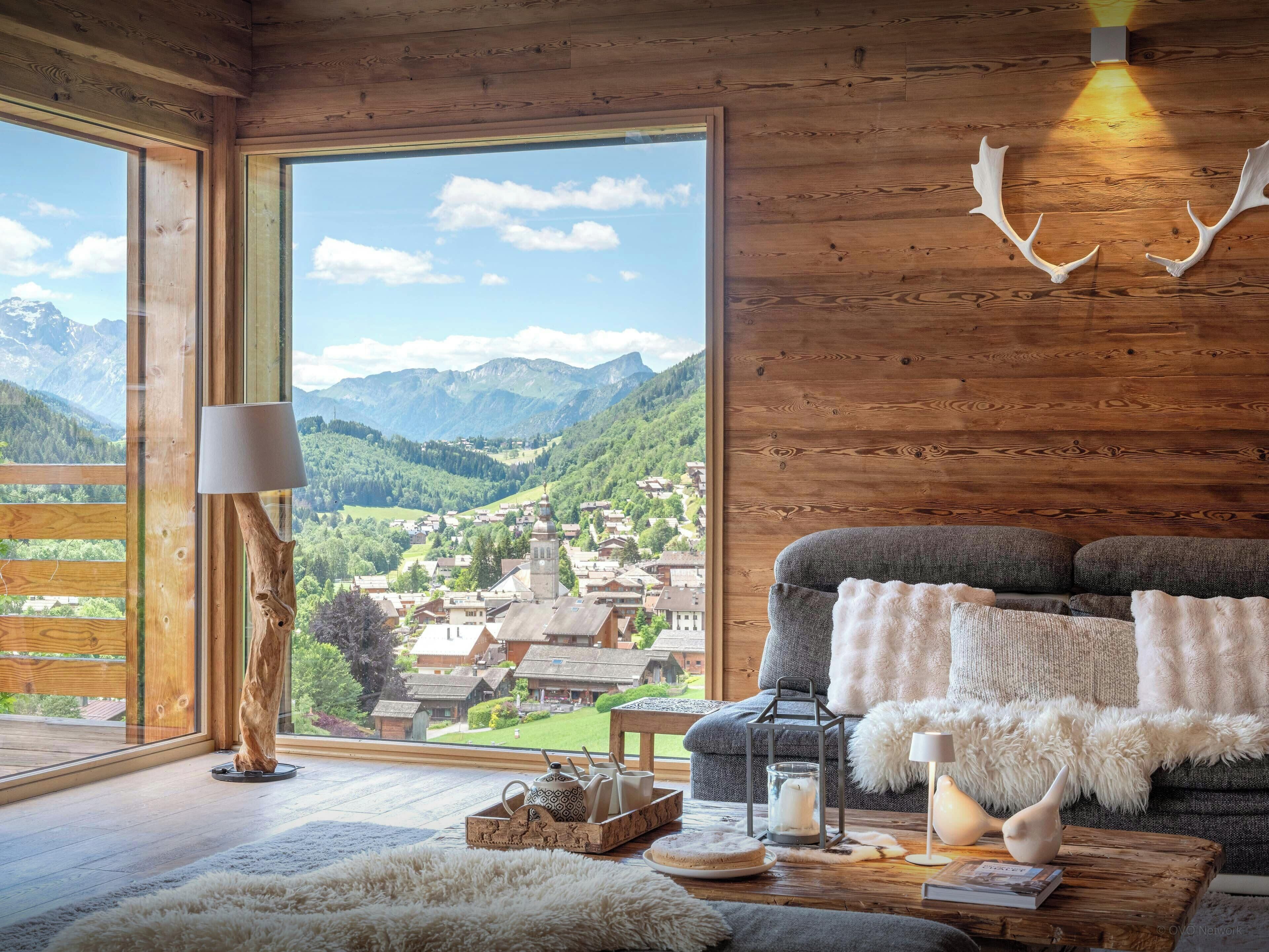 A cozy room with a large window, wooden walls, a grey sofa, a lamp, a rustic table, and a view of a village and mountains.