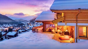 A wooden chalet with a balcony overlooking a snow-covered village and mountains.