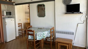 A compact kitchen with a refrigerator, microwave, and a small TV on a shelf. There is a dining table with chairs and a clock on the wall. - Cozy 4-Sleeper with A/C, Terraces & Direct Marina Access (Port Camargue)