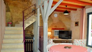 Una sala de estar acogedora con pared de piedra, una escalera, un televisor, una mesa de comedor con sillas y una pequeña cocina.
