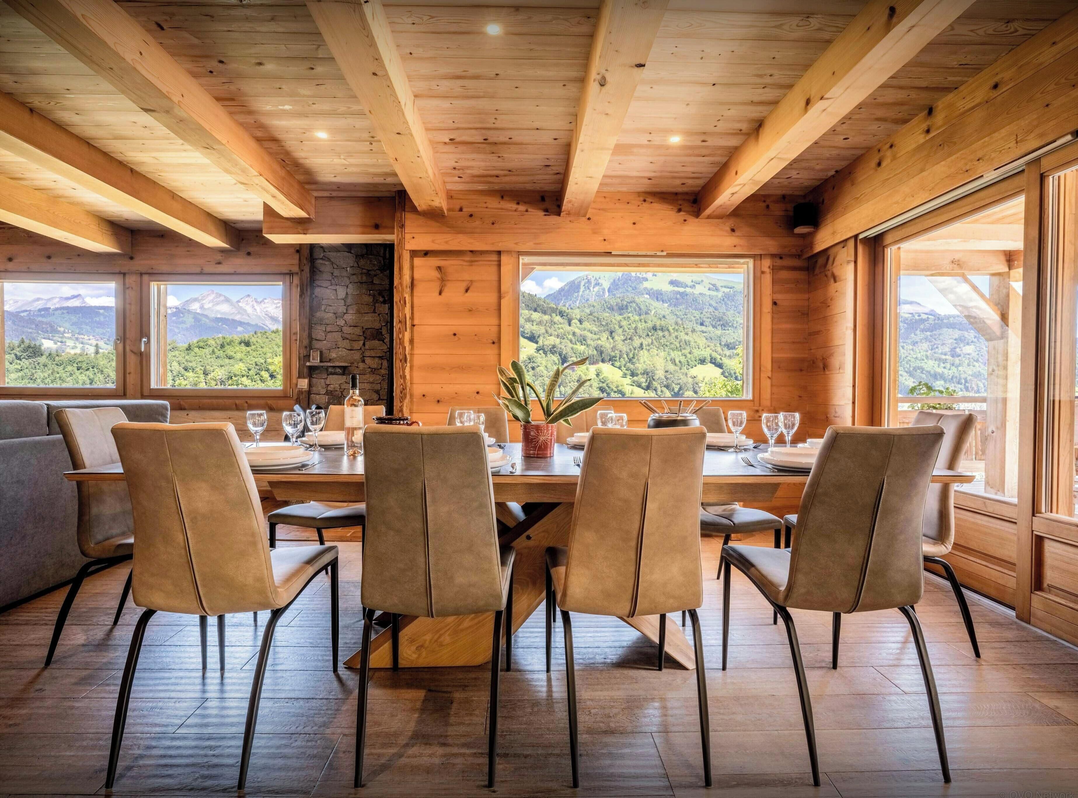 Ein Esszimmer mit Holzboden, einem Tisch für vier Personen, großen Fenstern mit Blick auf die Berge und einem Kamin aus Stein.