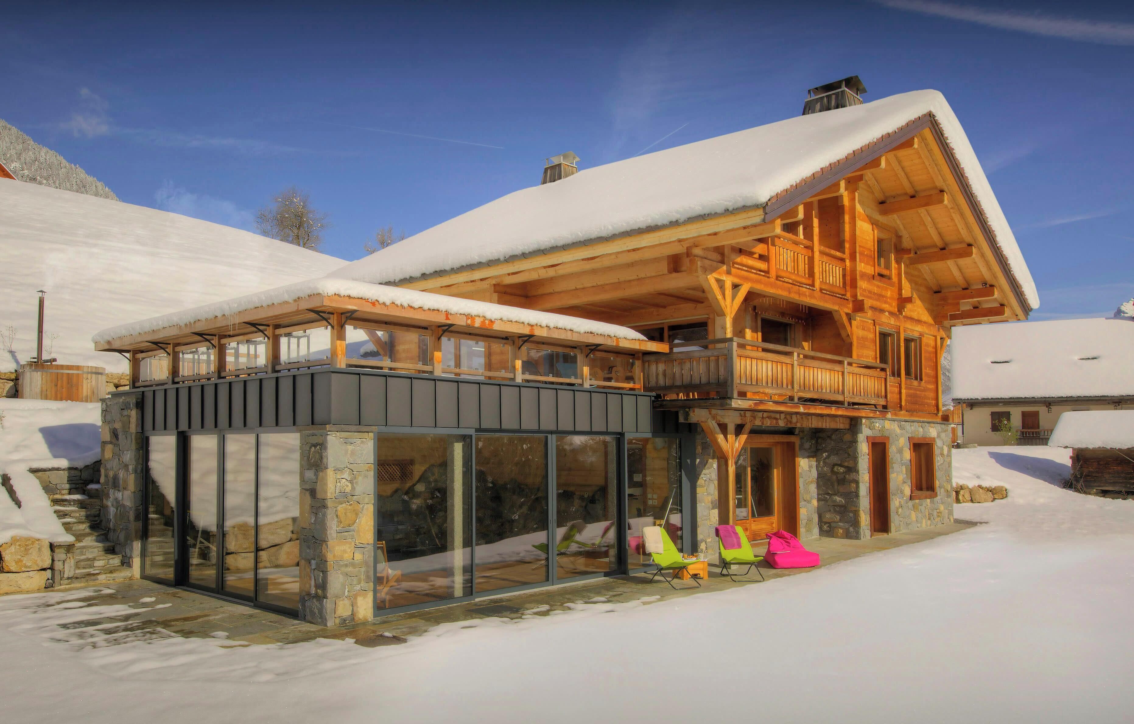 A wooden chalet with a balcony, stone walls, and a snow-covered roof.