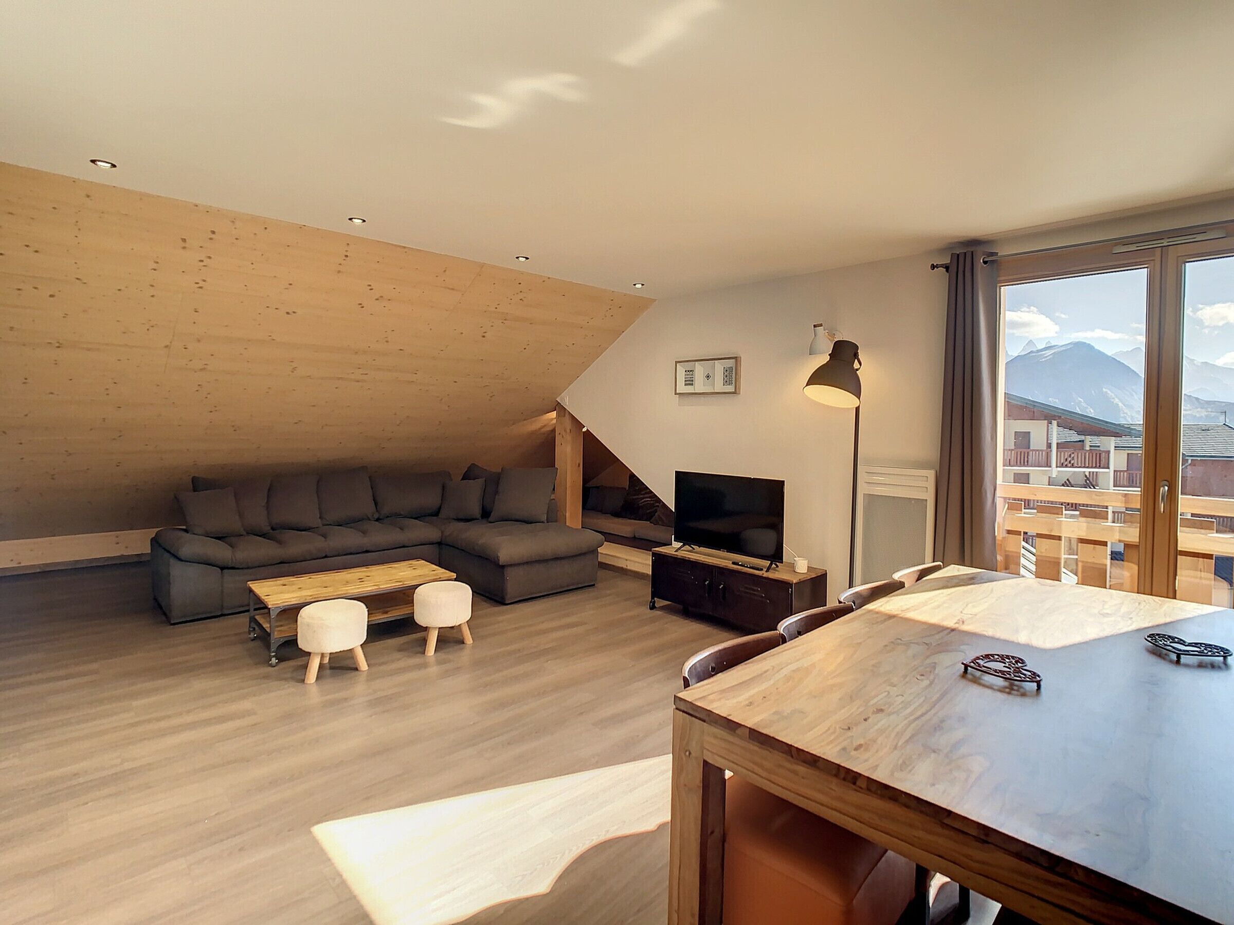 Ein stilvolles Loft-Wohnzimmer mit Holzbalkendecke, einem grauen Ecksofa, einem Couchtisch aus Holz und einem Esstisch mit Stühlen. Ein Fernseher steht auf einem Holzgestell, eine Stehlampe ist sichtbar.