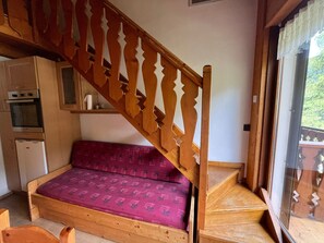 Een compacte kamer met een houten trap, een kleine kitchenette, een bed met een gestreept matras en uitzicht door een glazen deur.
