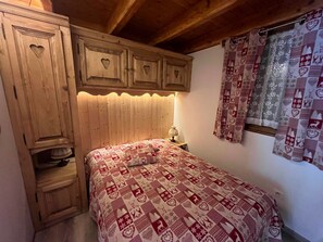 Een slaapkamer met een houten hoofdbord, een gebogen beddeken, een nachtkastje met een lamp en een houten garderobe met hartvormige uitsparingen.