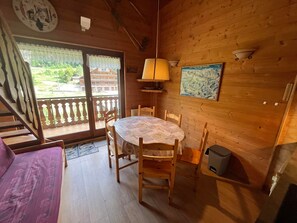 L’intérieur d’un chalet en bois, avec une table à manger, des chaises, une lampe, un tableau et une vue à travers une porte vitrée.