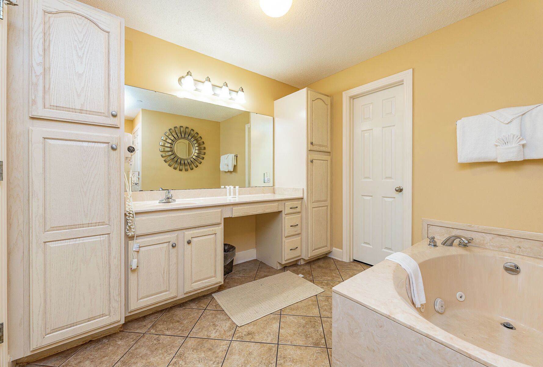 A bathroom with a vanity, a bathtub, a mirror with lights, a towel rack, and a door.