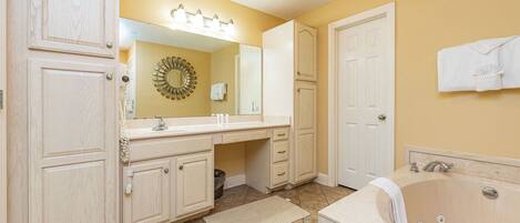 A bathroom with a vanity, a bathtub, a mirror with lights, a towel rack, and a door.