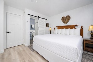 A hotel room with a bed, bedside lamps, a wooden headboard, a heart-shaped wall decoration, and a door.