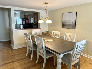 Un comedor con una mesa de madera y sillas blancas, una cocina al fondo y un cuadro en la pared.