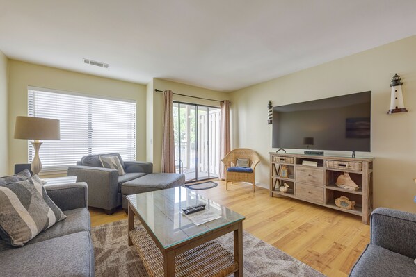 Una sala de estar con una mesa de centro de vidrio, sofás grises, un sillón de mimbre, un televisor sobre un mueble de madera y un ventanal con persianas.