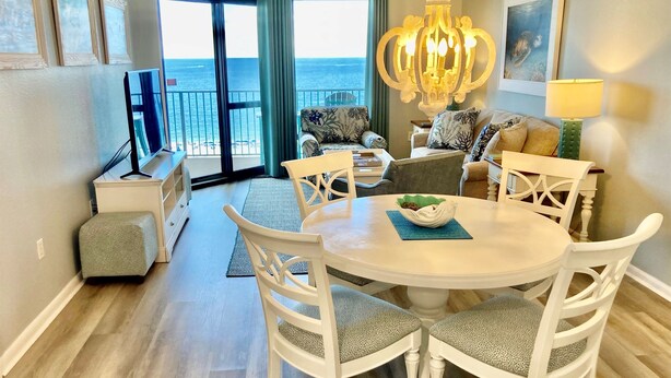 A dining area with a round table, white chairs, and a chandelier. There is a view of the ocean through large windows. - Gorgeous Gulf Views From This Orange Beach Condo on the 11th Floor! (Orange Beach)