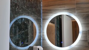 A bathroom with a marble wall, wooden floor, and two round mirrors with illuminated frames. - Hotel Havana (Sarandë)