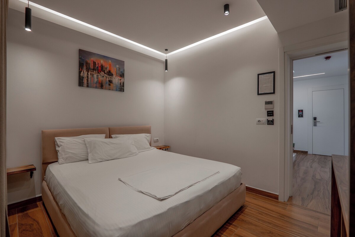 A modern bedroom with a large bed, a nightstand, a wall-mounted painting, and a door leading to another room.