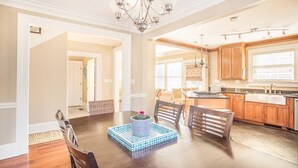 A spacious dining area with a wooden table and chairs, a chandelier, and a kitchen with wooden cabinets and a window. - Downtown Historic District Gem w/ Courtyard + 3 King Beds by Lucky Savannah (Savannah)